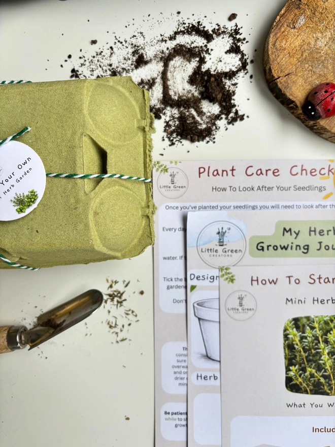 green egg box tied with green thread and a grow your own mini herb garden tag, Placed on a white background with instructions next to it plus a wooden disc, spilled soil and a gardening tool