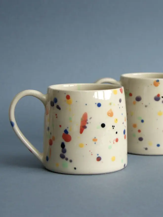 rainbow splatter hand-thrown ceramic mug. pair of mugs photographed against blue studio background.