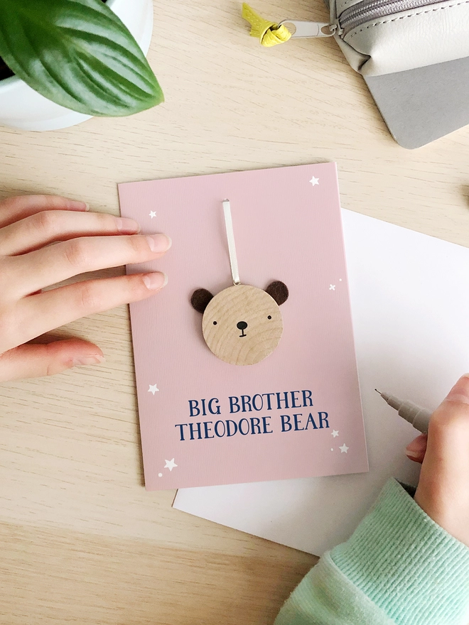 A pink greetings card with a small wooden bear keepsake and the words "Big brother Theodore bear" printed on is on a wooden desk. 