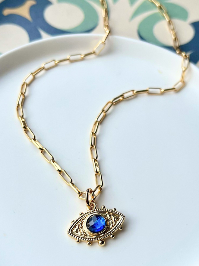 Blue evil eye charm with gold paperclip chain on white dish