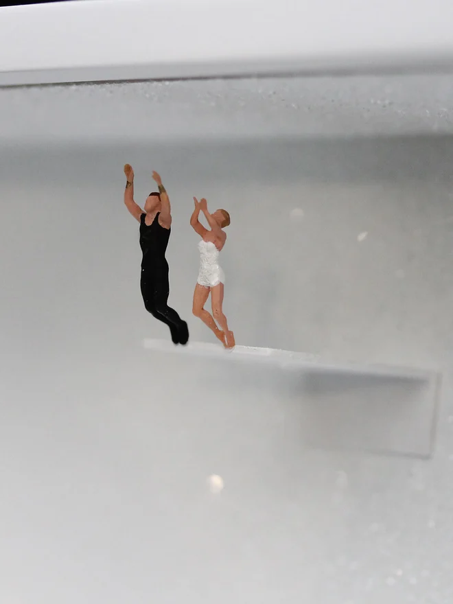 Miniature scene in an artbox showing a tiny woman and man about to dive off a diving board, set against a sparkling white backdrop (close up). 