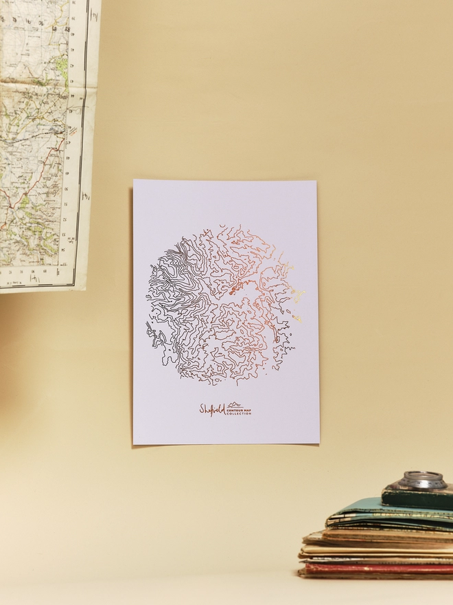 Styled Sheffield metallic copper contour map print with stack of maps in the foreground and a compass