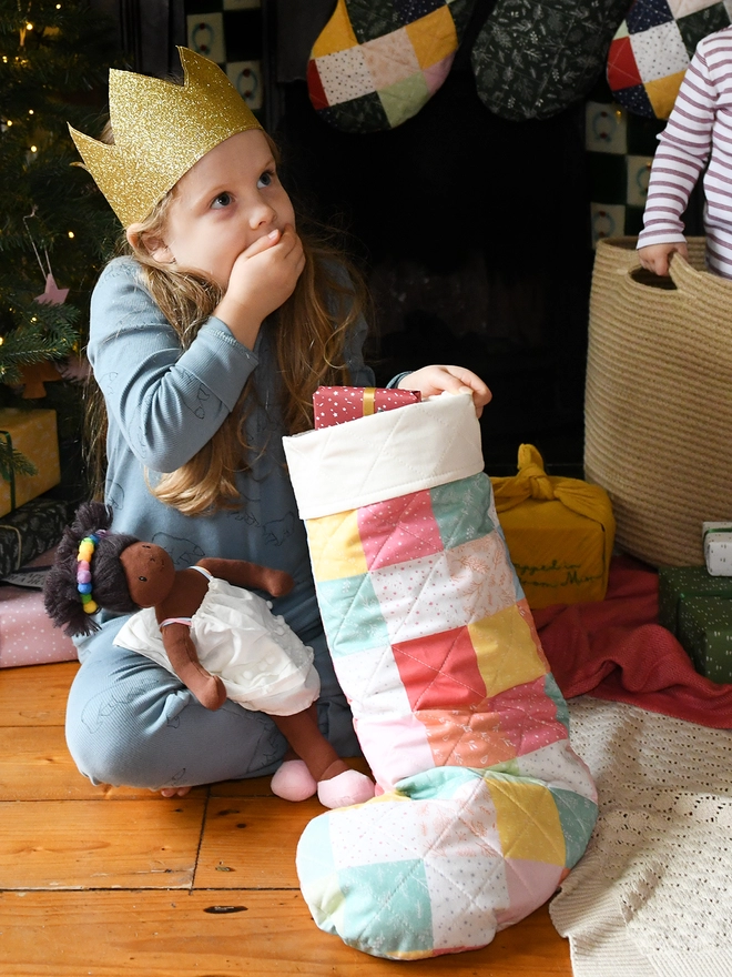 A young girl wearing blue pyjamas and a golden crown sits in front of a fireplace and holds a pastel patchwork Christmas stocking in her arms.