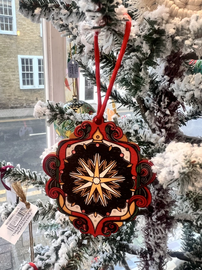 Multi coloured wooden tree decoration with a star pattern at the centre. It is hanging by a red ribbon on a fake white christmas tree with other decorations. 