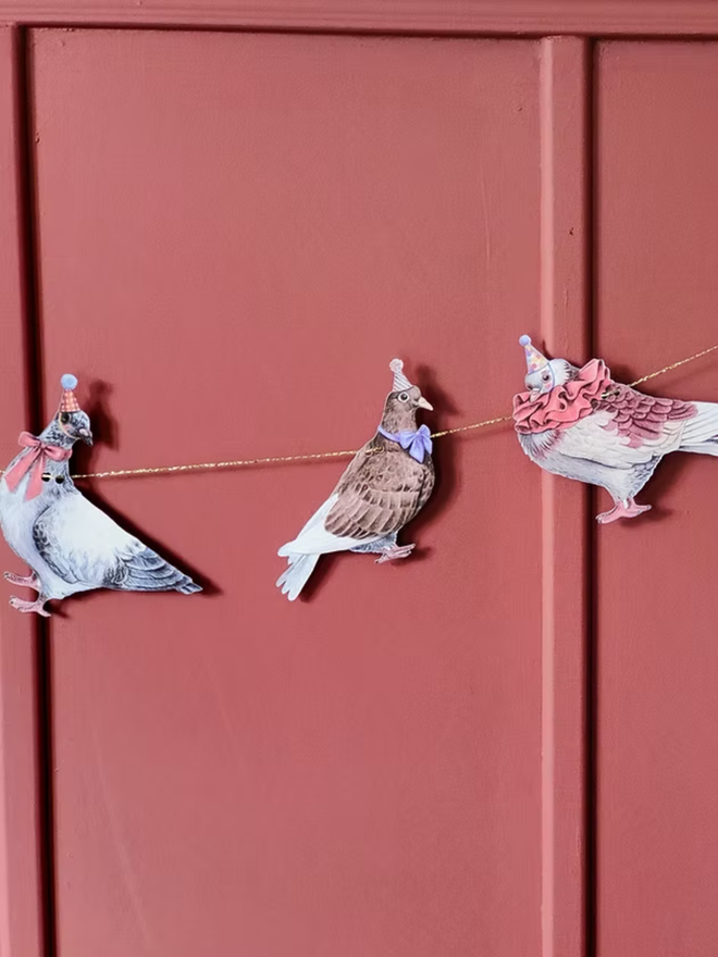 party pigeons circus garland. a string of paper pigeons in party and circus hats hung up on a wall.