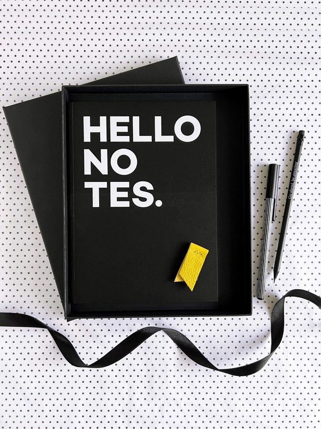 Open black gift box with HELLO NOTES notebook, yellow leather marker, pencil and pen on spotty paper