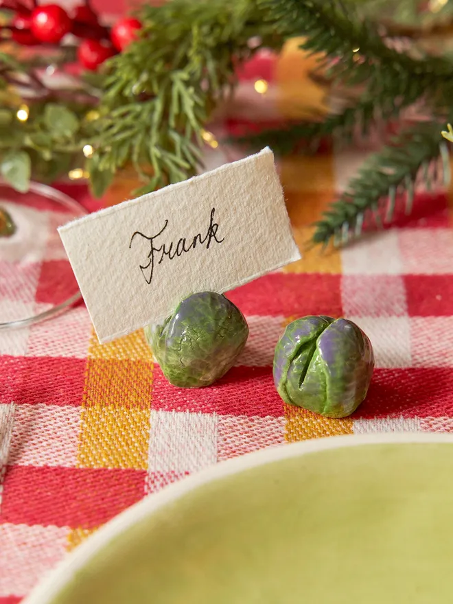 A set of handmade ceramic Brussels sprout name card holders arranged on a yellow and red checkered tablecloth. Each glossy green sprout has individually modelled leafy details, and one is shown holding a heart-shaped display card.