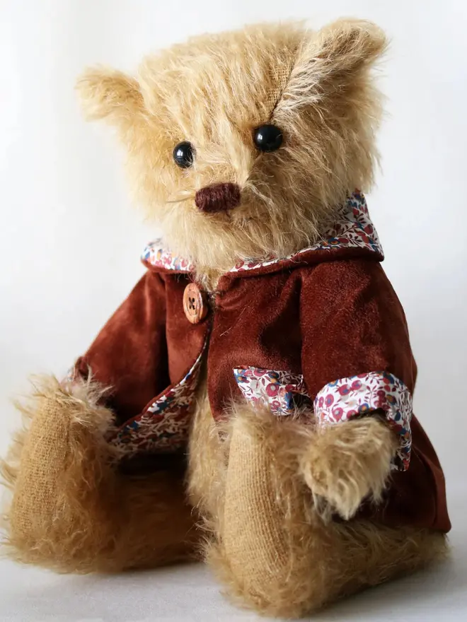 Brown teddy bear wearing a burgundy cloak with flower details and a brown button, sitting upright against a white background.