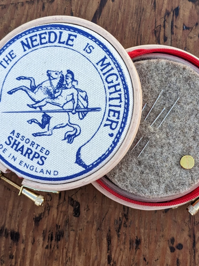 Royal blue coloured 'the needle is mightier' embroidery hoop needle book depicting a soldier on horse back with a needle as a sword shown unzipped with inner felt and needles