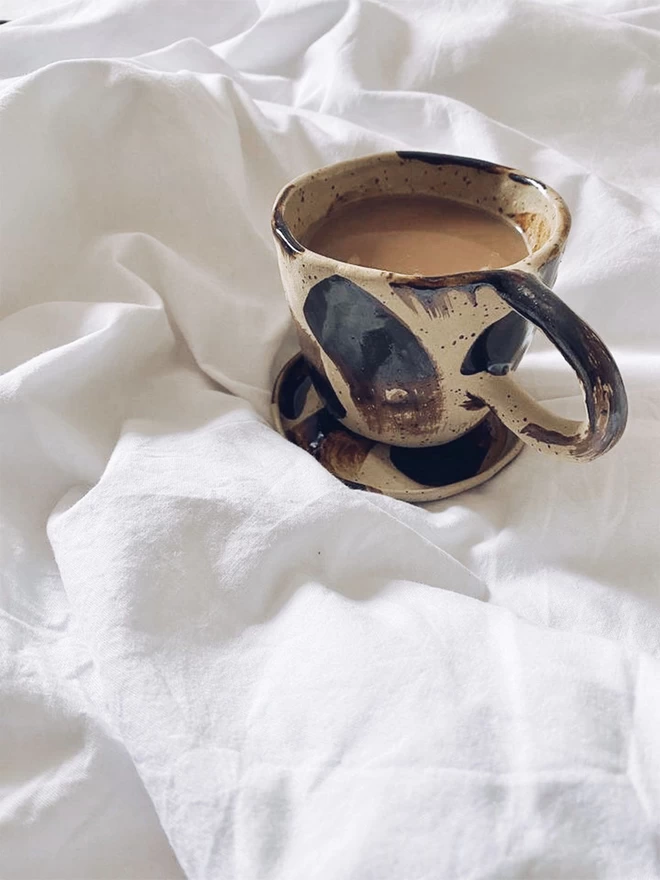 beige ceramic mug and saucer with a black brushstroke design, sat on a bed