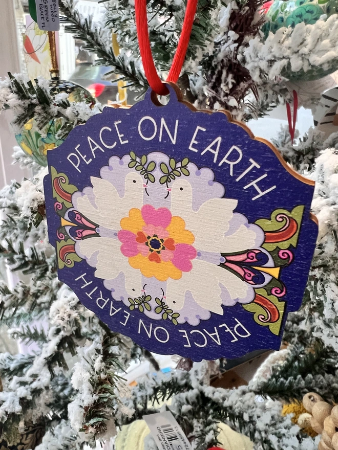 Multi coloured wooden tree decoration the reads 'Peace on Earth' around it, with four white doves and a flower pattern at the centre. It is hanging by a red ribbon on a fake white christmas tree with other decorations. 