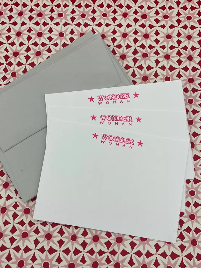South London Letterpress Wonder Woman notecards seen with grey envelopes on a patterned pink and red background.
