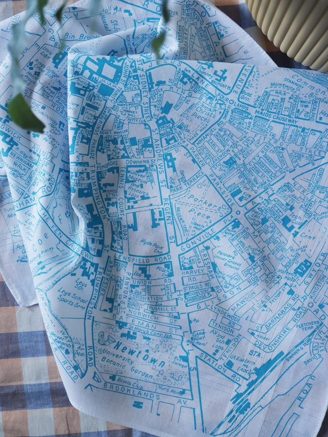 A lifestyle photo of a Mr.PS Cambridge Map hankie printed in aqua with a plant pot on a gingham tablecloth
