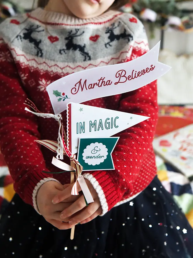 A child wearing a traditional red Christmas jumper holds three personalised Christmas flags made from cardboard. Each flag is attached to a wooden stick with ribbons. The flags read "Martha Believes in magic & wonder".