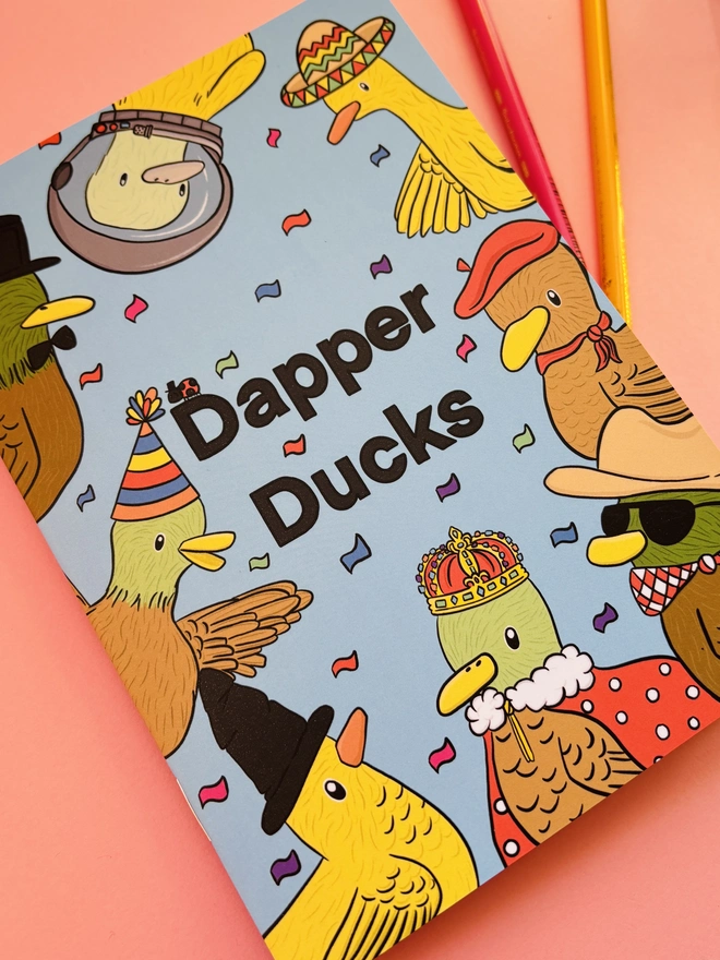 Dapper Ducks colouring book shows an A5 book of lots of illustrated ducks in different hats including a crown, beret, party hat and sombrero. A full colour cover reads “Dapper ducks” in black on a blue background.