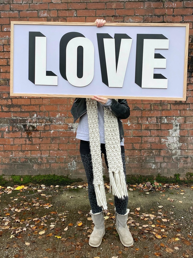 A person standing in front of a red brick wall holding a painted wooden sign which reads LOVE on a lilac painted back board 