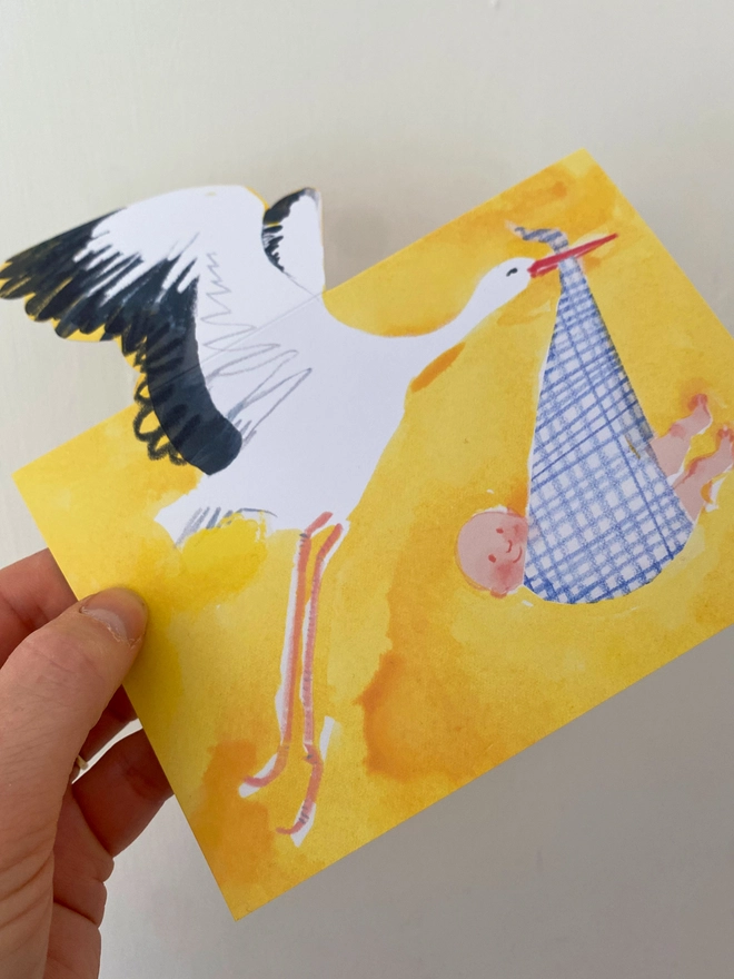 A new baby card by Esther Kent showing a black and white stork carrying a baby in a blue checked cloth, on a yellow background
