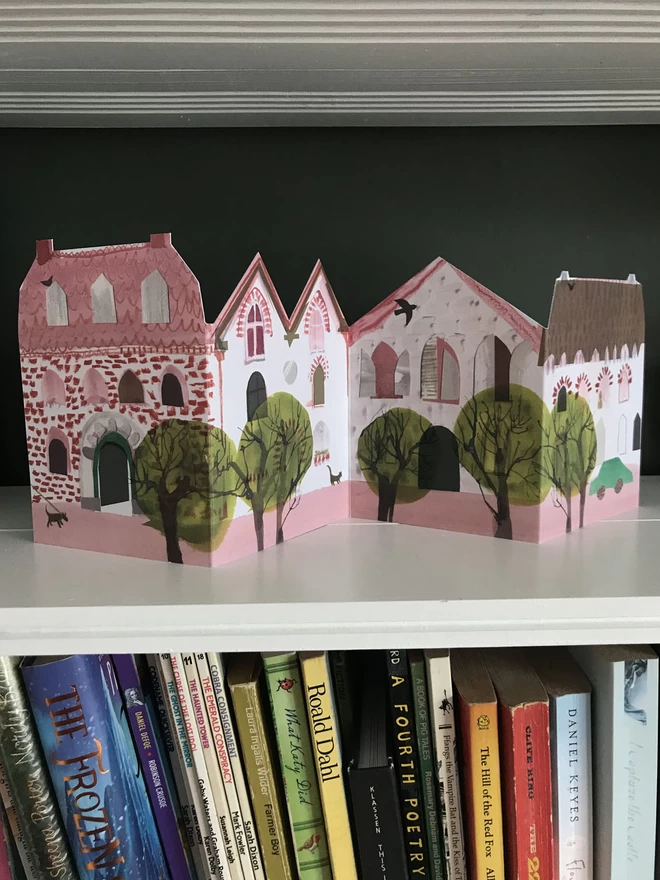 Folded concertina greetings card showing pink houses sits on a shelf above books, against a dark green background