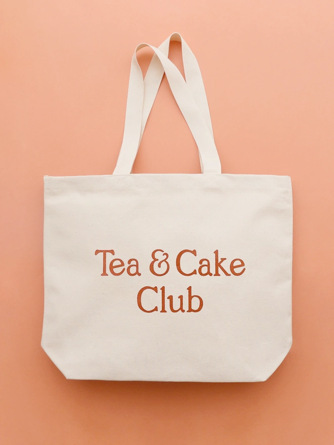 a large natural canvas tote bag bearing the words tea and cake club laying on an orange backdrop