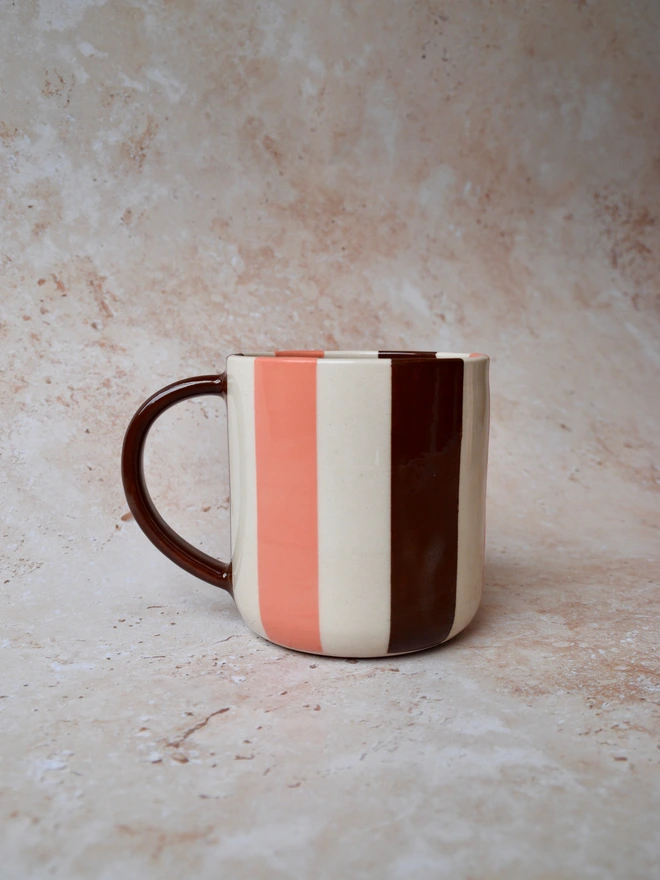 Handmade striped ceramic mug with vertical chocolate brown and peach orange stripes on a creamy white background, featuring a dark brown handle and photographed on a warm neutral textured backdrop.