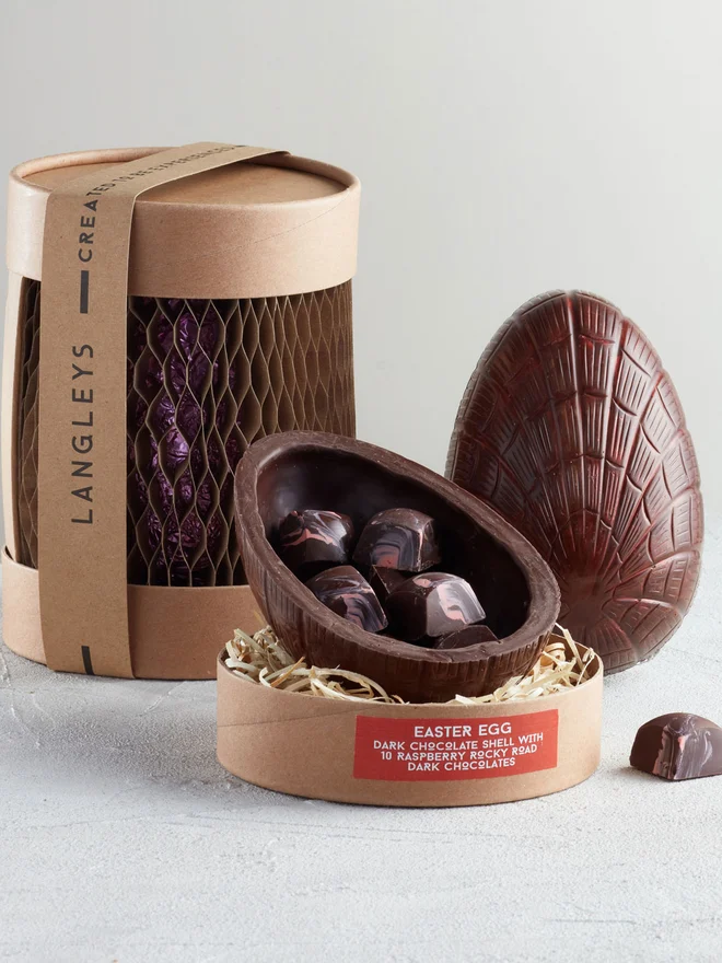 Dark Chocolate Easter Egg With Raspberry Rocky Road, a chocolate egg sitting on a white surface. It is accompanied by another chocolate egg that is opened showing the chocolates within. 