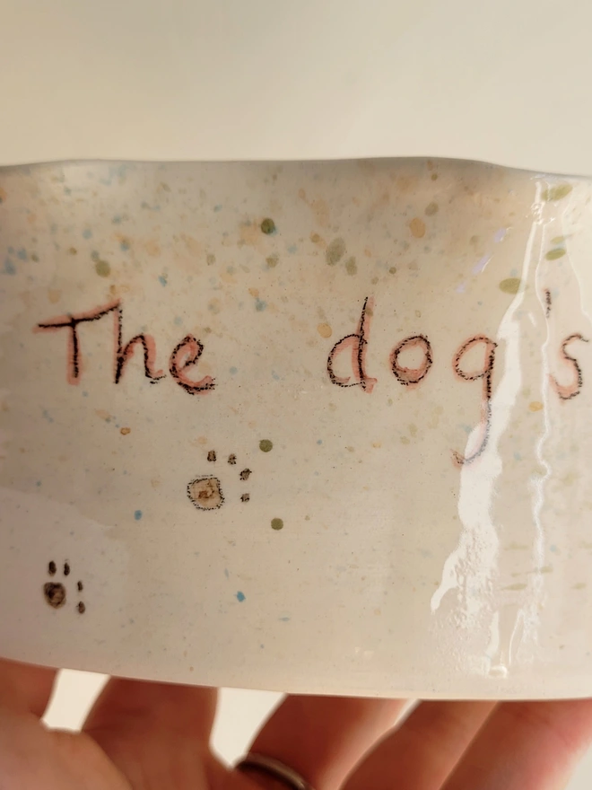 ivory ceramic dog bowl with 'the dog's dinner' written on the side with brown green and blue splatters and pawprints held in a hand