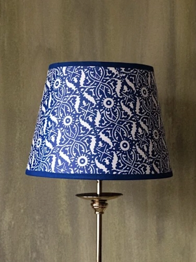 Retro Drum Lampshade. A tapered drum lampshade covered in a bold blue and white ornamental block print with a matching blue trim, shown on a brass lamp base. The striking patterned shade brings classic blue and white style to a sitting room or bedroom.