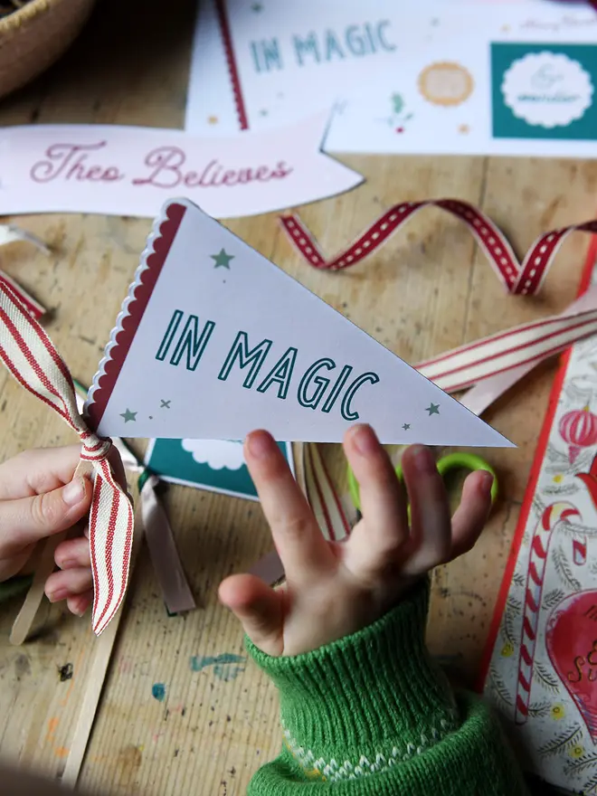 A young child holds a Christmas flag that reads "in magic" above a wooden table covered in various craft items.