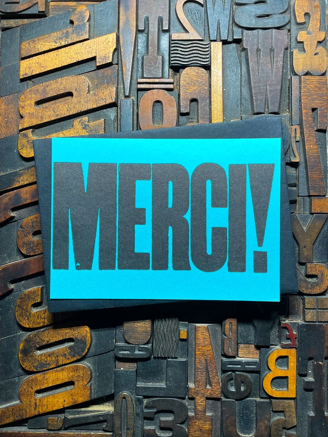 A bright turquoise thick duplex card with the word "MERCI!" printed in bold, black letters lies on a surface covered with various letterpress blocks.