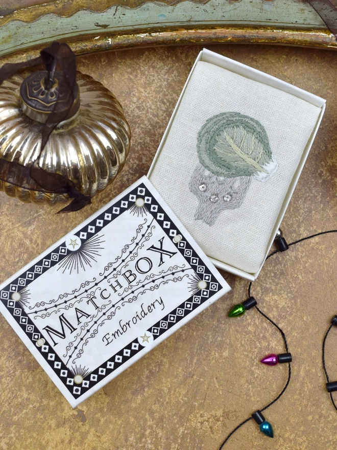 The Brussels Sprout embroidery, inside a Matchbox displayed on a gold background with glass baubles.