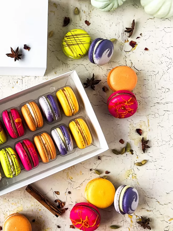 an assortment of colourful autumn macarons are arranged in a white box, surrounded by mixed autumn spices