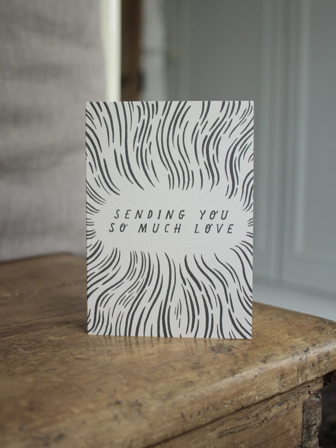 Black and white greeting card with illustration and the words sending you so much love on it stood up on a wooden surface