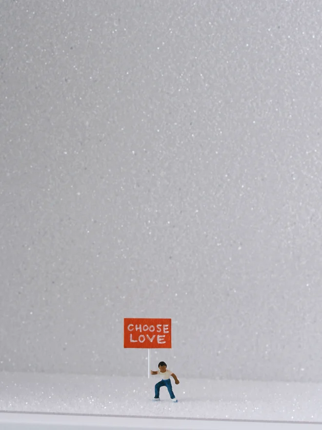 Miniature scene in an artbox showing a tiny child is holding a protest sign saying "Choose Love" against a sparkling white backdrop (close up)