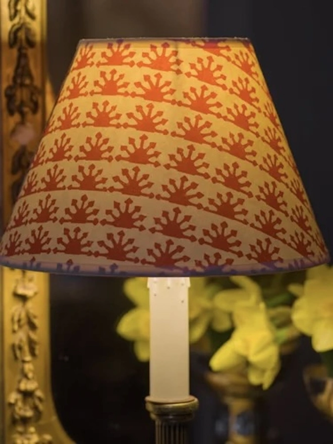 Paper Empire Lampshade. A glowing lampshade printed with a repeating red sunburst pattern, lit on a candlestick lamp beside yellow daffodils and a gilt mirror. The illuminated block-print shade casts a warm, inviting light for cosy evening interiors.