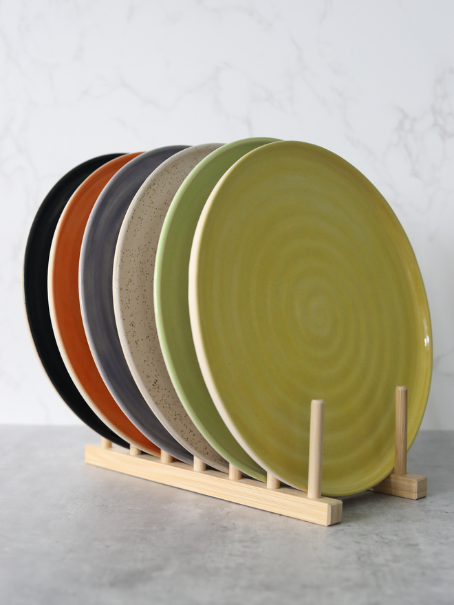Six handmade ceramic dinner plates displayed upright in a natural wood plate rack, fanned out to reveal a colour palette of lime green, sage, speckled stone, slate grey, terracotta, and matte black, against a white marble background