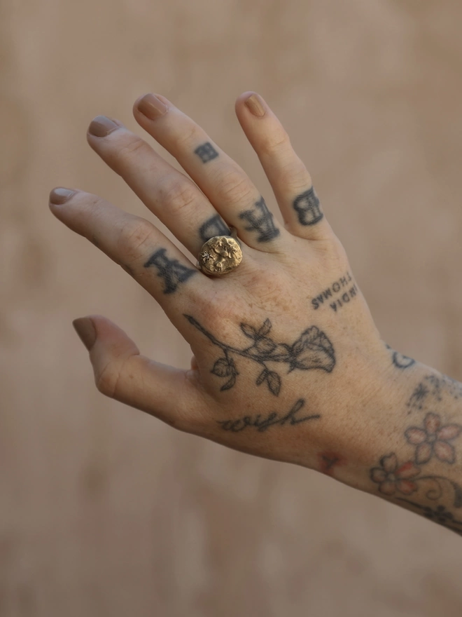 Image of a tattooed female hand wearing a gold coloured brass ring with the image of a horse and star on it. The background of the image is a terracotta coloured rough plaster 