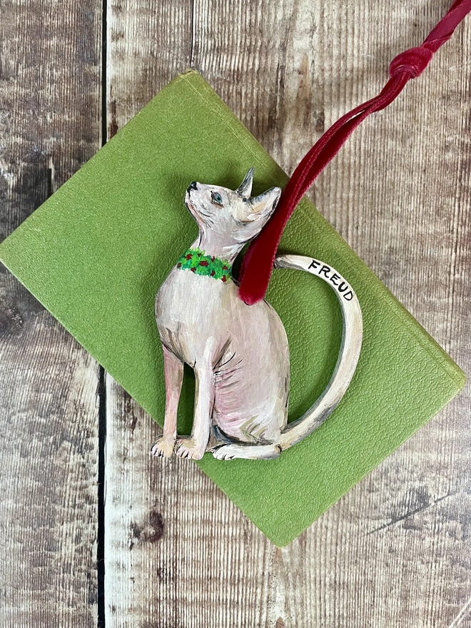 Hairless Sphynx Cat Christmas Decoration strung with red velvet ribbon and placed on a green book
