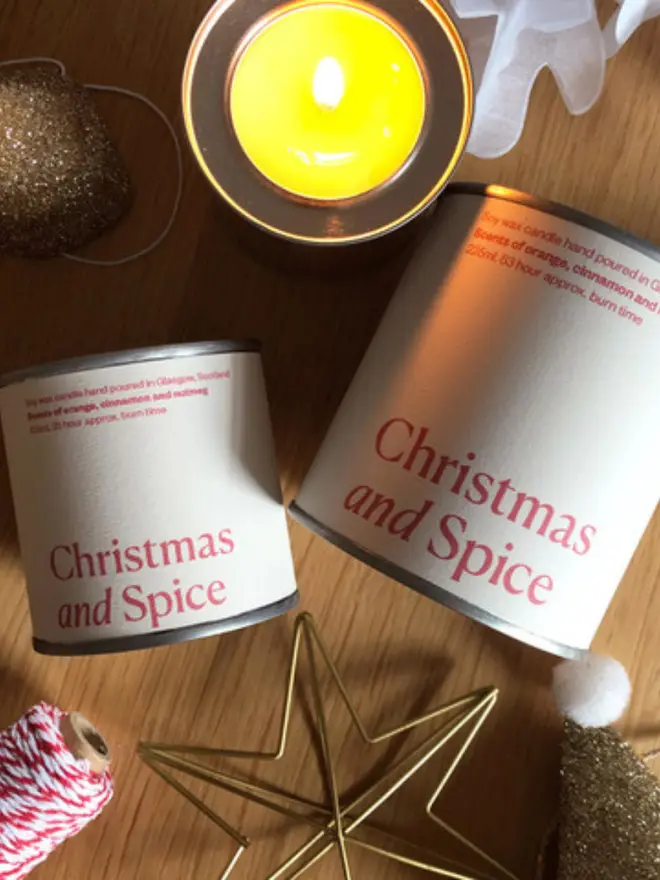 Christmas and Spice Wax Candle - a pale pink and red tin in a small and big size held up against some christmas items 