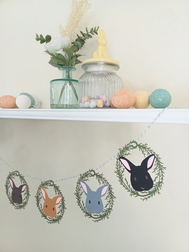 6 Bunny Easter Garland strung across shelf. Each bunny face displayed inside oval green lasercut foliage wreath. Left to righy: Dark Brown, Toffee, Dark Grey, Black bunny face all with pink ears