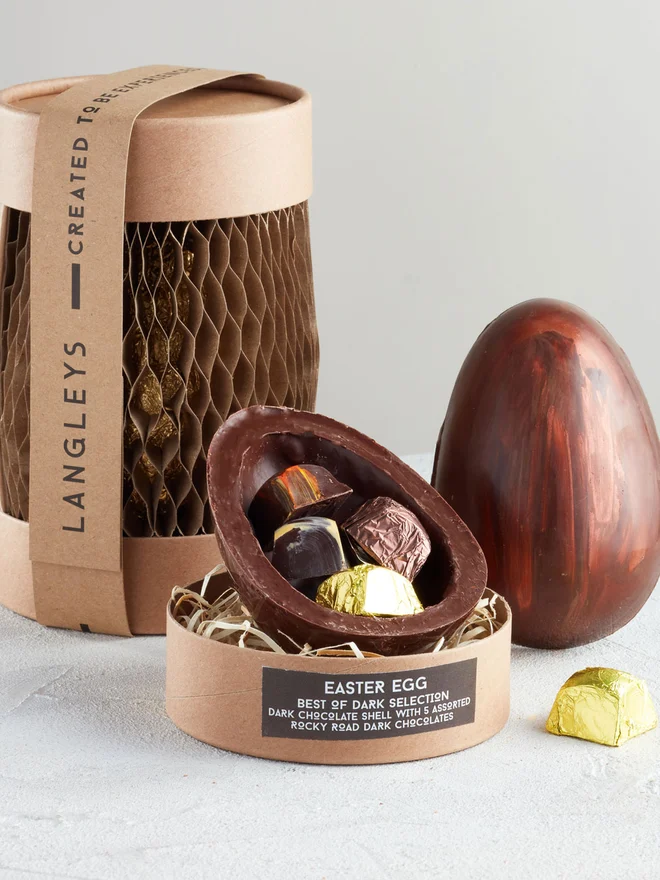 Best Of Dark Chocolate Easter Egg, a chocolate egg sitting on a white surface. It is accompanied by another chocolate egg that is opened showing the chocolates within. 