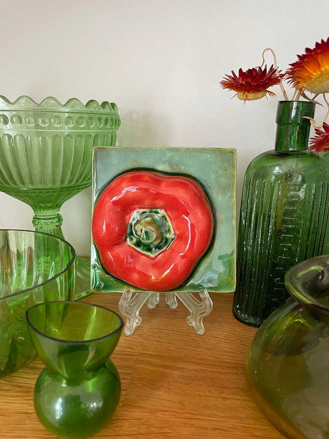 Decorative eye-catching red pepper feature tile – three-dimensional, very realistic and incredibly tactile - displayed on a mantelpiece alongside antique green glass.   