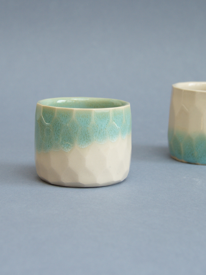 Two straight sided cups, one in the centre of the image and one half in view in the background.  The central one has a turquoise top half and a cream bottom half while the other one has the opposite.  The surface of the cup has a hexagonal texture.
