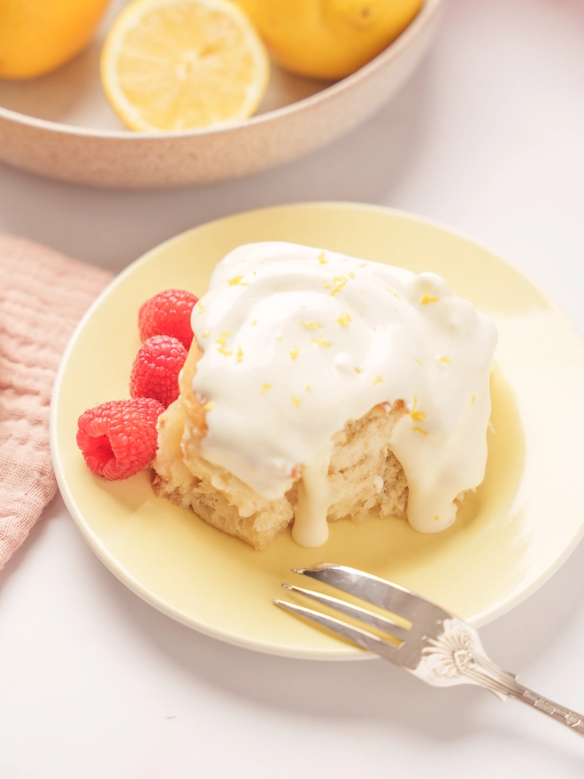 Lemon Drizzle Bun on a plate with 3 raspberries, with a fork and a pink bowl of lemons in the background