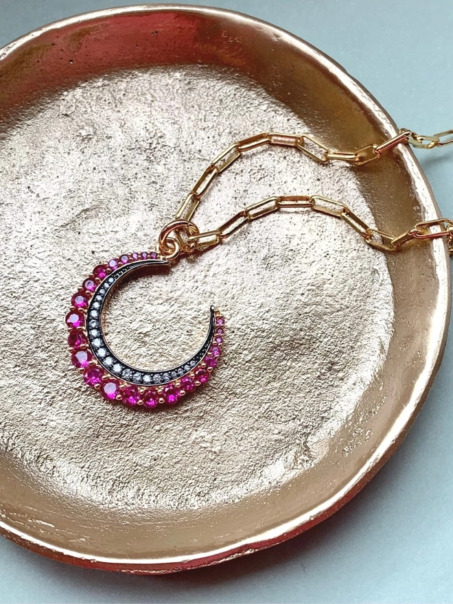 Pink jewelled crescent moon charm on a gold paperclip chain inside a small gold bowl on a green background