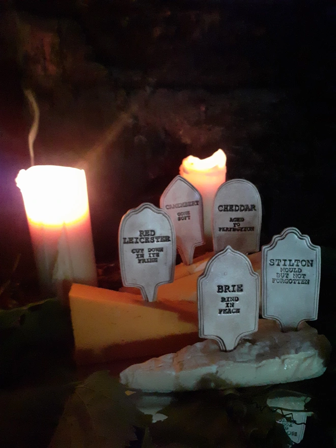 Five ceramic gravestone cheese markers arranged among wedges of cheese in a dark, candlelit setting, with smoke curling above a glowing candle.