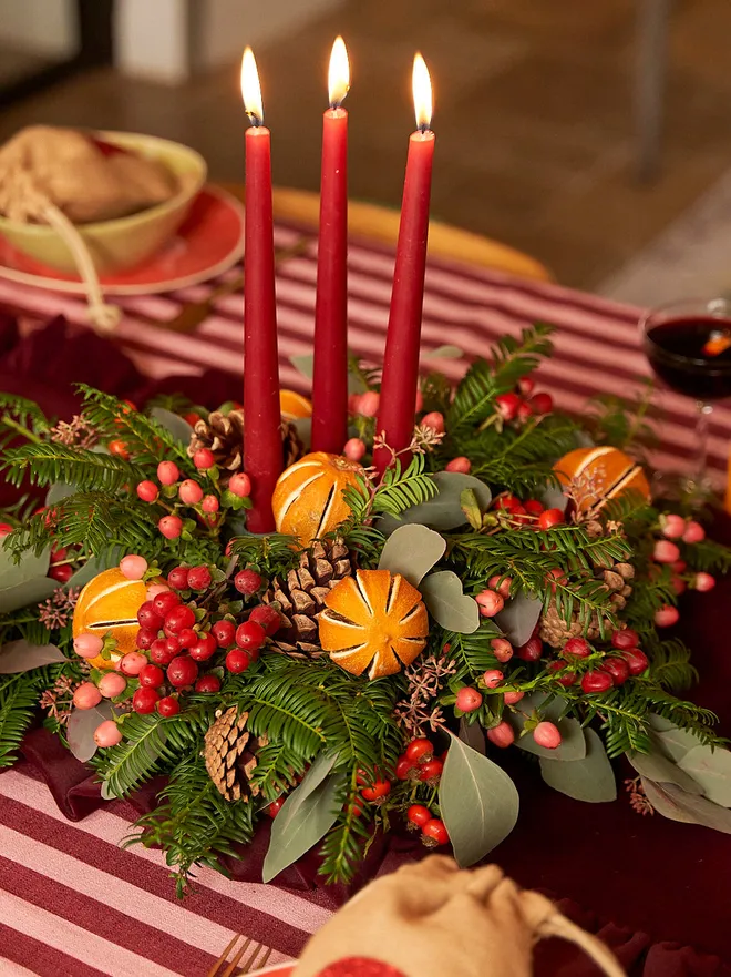 green wreath with dried oranges, red berries, and leaves on a table with red striped table cloth. three lit, red candles in the middle