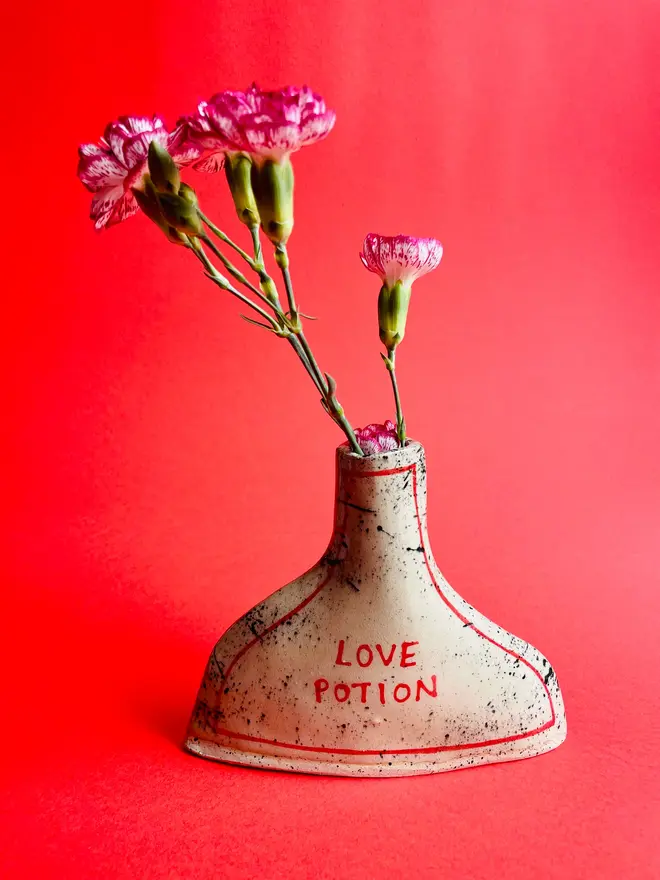 Love Potion/ Poison Bottle Vase, a vase that says 'love potion' in front of a red backdrop. Each is stamped with a signature red dot on the bottom. 