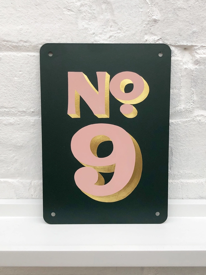 Hand painted house number No. 9 in pale pink and gold leaf on anthracite grey metal plaque, against a white brick wall. 