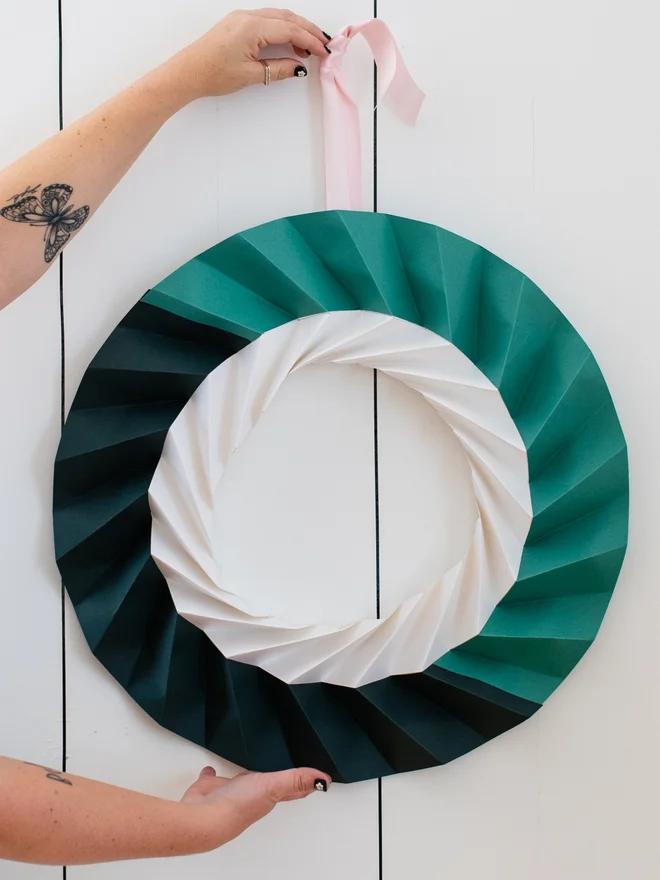 Woman's arm stretched out with a modern origami wreath hanging in dark green, jade green and white, hung from pale pink ribbon