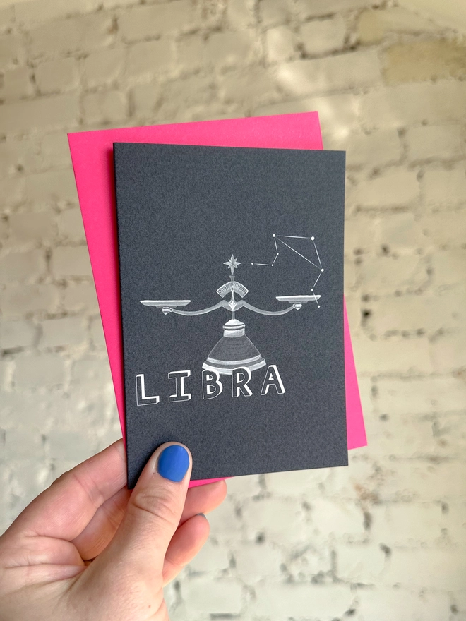  a greetings card featuring a greyscale illustration of a zodiac style set of scales with the text “LIBRA” with a neon pink envelope